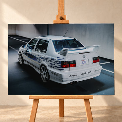 Weißer VW Jetta in rückwärtiger Dreiviertelansicht in einem Parkbereich, mit blauen Flammengrafiken, großem Heckflügel, hellen Mehrspeichenrädern und roten Rückleuchten bei kühlem Licht; Visualisierung als Leinwand-Wandbild, frontal auf einer Holzstaffelei.