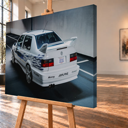 Weißer VW Jetta in rückwärtiger Dreiviertelansicht in einem Parkbereich, mit blauen Flammengrafiken, großem Heckflügel, hellen Mehrspeichenrädern und roten Rückleuchten bei kühlem Licht; Visualisierung als Leinwand-Wandbild, schräg von der Seite auf einer Holzstaffelei.
