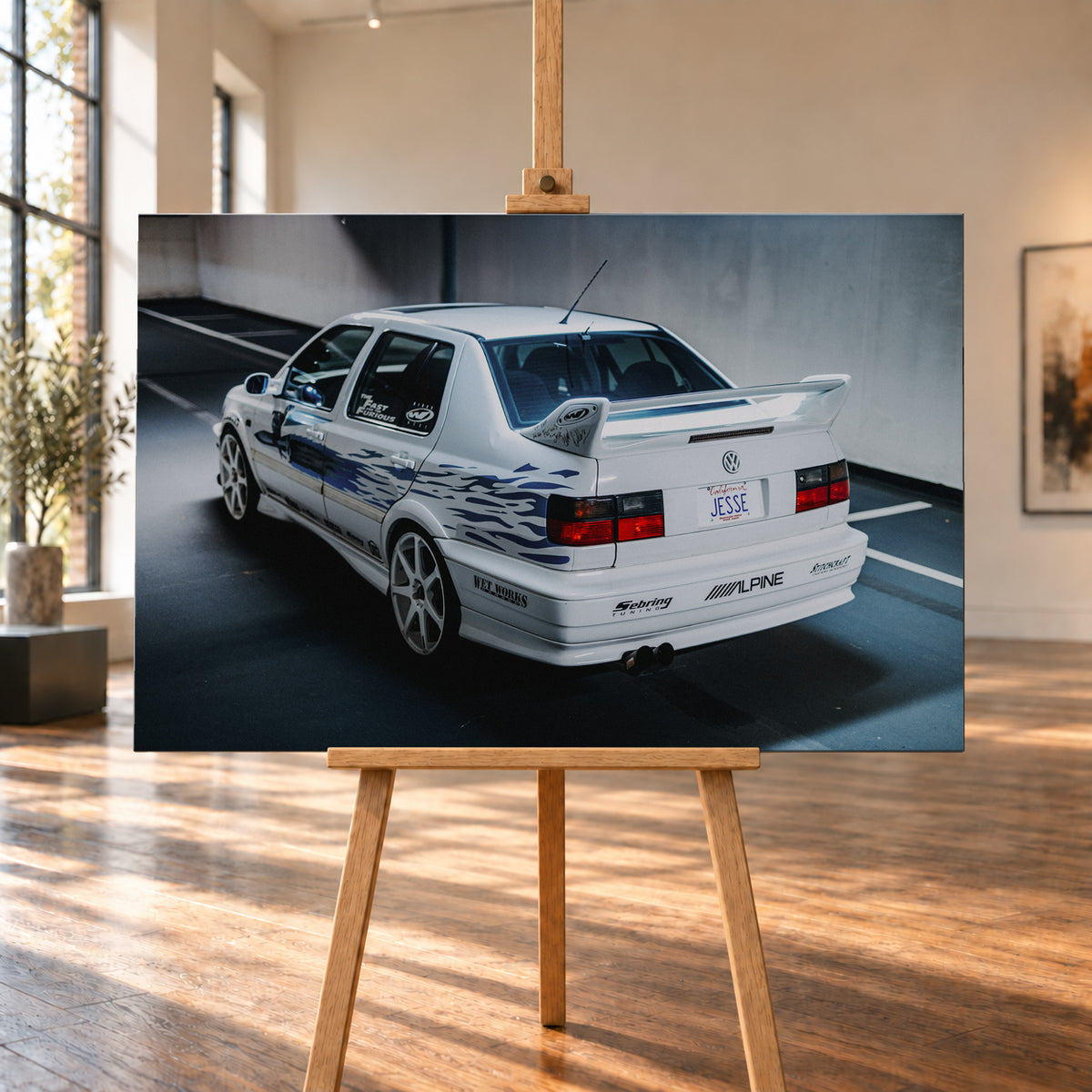 Weißer VW Jetta in rückwärtiger Dreiviertelansicht in einem Parkbereich, mit blauen Flammengrafiken, großem Heckflügel, hellen Mehrspeichenrädern und roten Rückleuchten bei kühlem Licht; Visualisierung als Leinwand-Wandbild, auf einer Holzstaffelei in einem hellen Galerieraum.