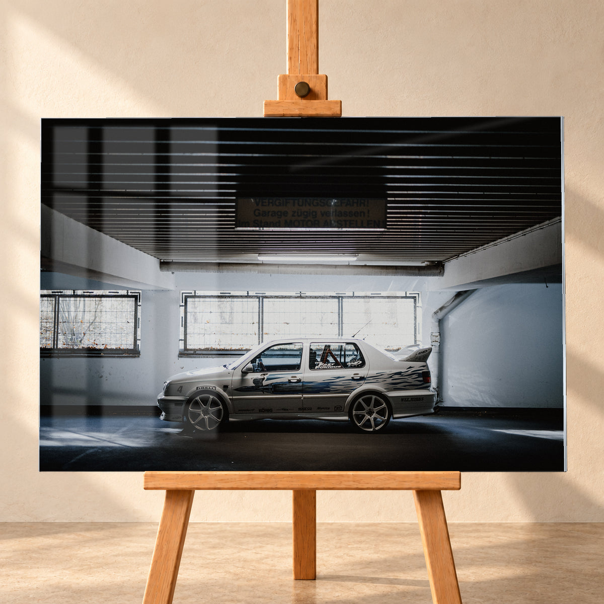 Silberfarbener VW Jetta mit blau-schwarzer Seitengrafik, großen Leichtmetallrädern und hohem Heckflügel im Seitenprofil in einer dunklen Parkgarage; Visualisierung als Acrylglas-Wandbild, frontal auf einer Holzstaffelei.