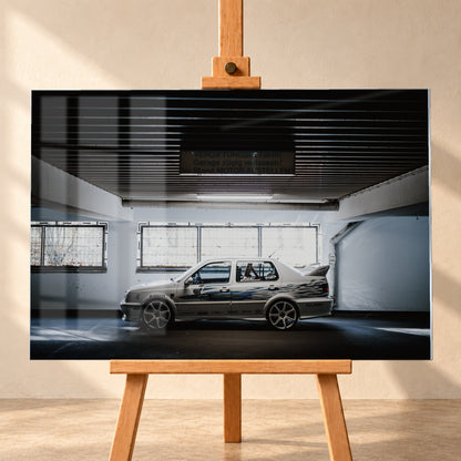 Silberfarbener VW Jetta mit blau-schwarzer Seitengrafik, großen Leichtmetallrädern und hohem Heckflügel im Seitenprofil in einer dunklen Parkgarage; Visualisierung als Acrylglas-Wandbild, frontal auf einer Holzstaffelei.