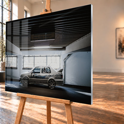 Silberfarbener VW Jetta mit blau-schwarzer Seitengrafik, großen Leichtmetallrädern und hohem Heckflügel im Seitenprofil in einer dunklen Parkgarage; Visualisierung als Acrylglas-Wandbild, schräg von der Seite auf einer Holzstaffelei.