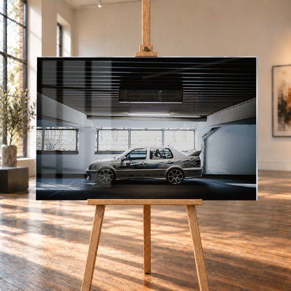 Silberfarbener VW Jetta mit blau-schwarzer Seitengrafik, großen Leichtmetallrädern und hohem Heckflügel im Seitenprofil in einer dunklen Parkgarage; Visualisierung als Acrylglas-Wandbild, auf einer Holzstaffelei in einem hellen Galerieraum.