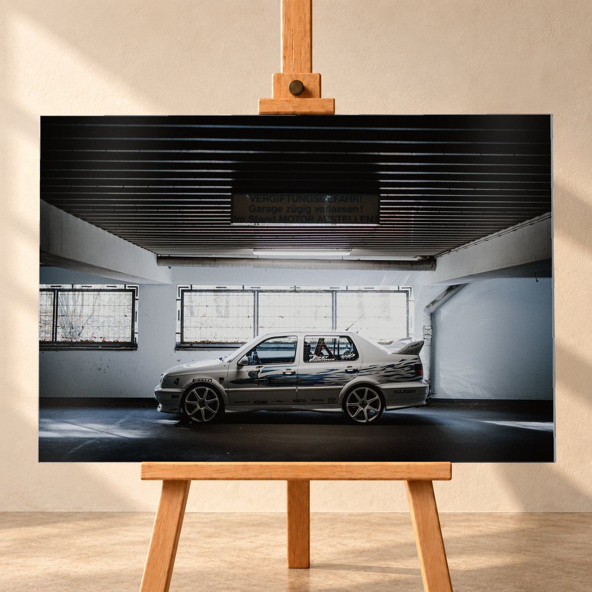 Silberfarbener VW Jetta mit blau-schwarzer Seitengrafik, großen Leichtmetallrädern und hohem Heckflügel im Seitenprofil in einer dunklen Parkgarage; Visualisierung als Alu gebürstet-Wandbild, frontal auf einer Holzstaffelei.