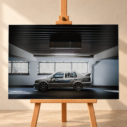 Silberfarbener VW Jetta mit blau-schwarzer Seitengrafik, großen Leichtmetallrädern und hohem Heckflügel im Seitenprofil in einer dunklen Parkgarage; Visualisierung als Alu gebürstet-Wandbild, frontal auf einer Holzstaffelei.