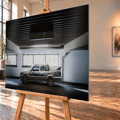 Silberfarbener VW Jetta mit blau-schwarzer Seitengrafik, großen Leichtmetallrädern und hohem Heckflügel im Seitenprofil in einer dunklen Parkgarage; Visualisierung als Alu gebürstet-Wandbild, schräg von der Seite auf einer Holzstaffelei.