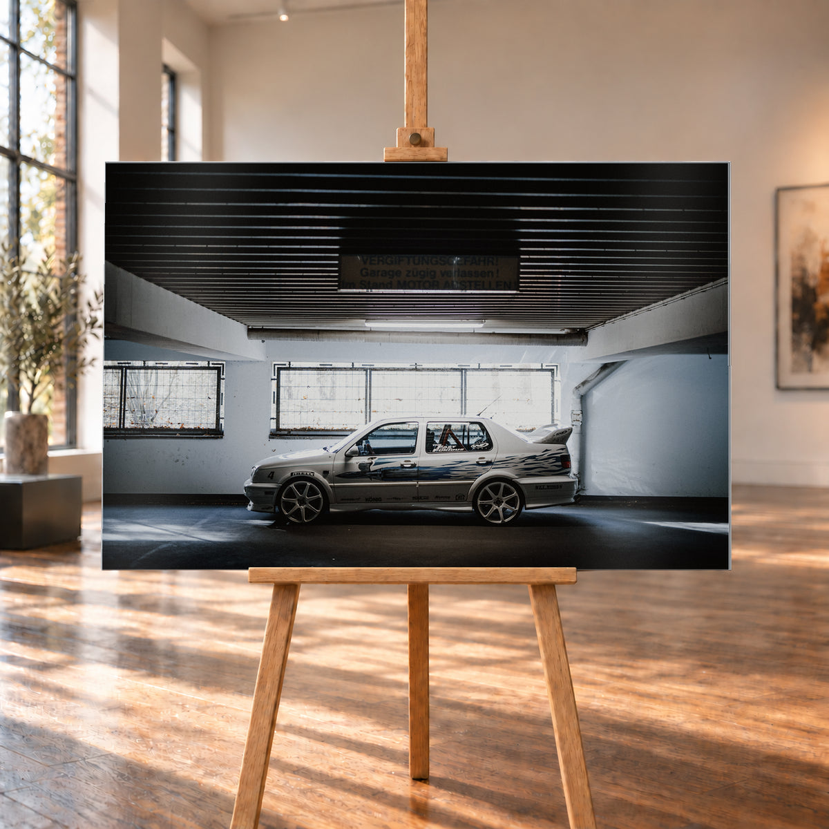 Silberfarbener VW Jetta mit blau-schwarzer Seitengrafik, großen Leichtmetallrädern und hohem Heckflügel im Seitenprofil in einer dunklen Parkgarage; Visualisierung als Alu gebürstet-Wandbild, auf einer Holzstaffelei in einem hellen Galerieraum.