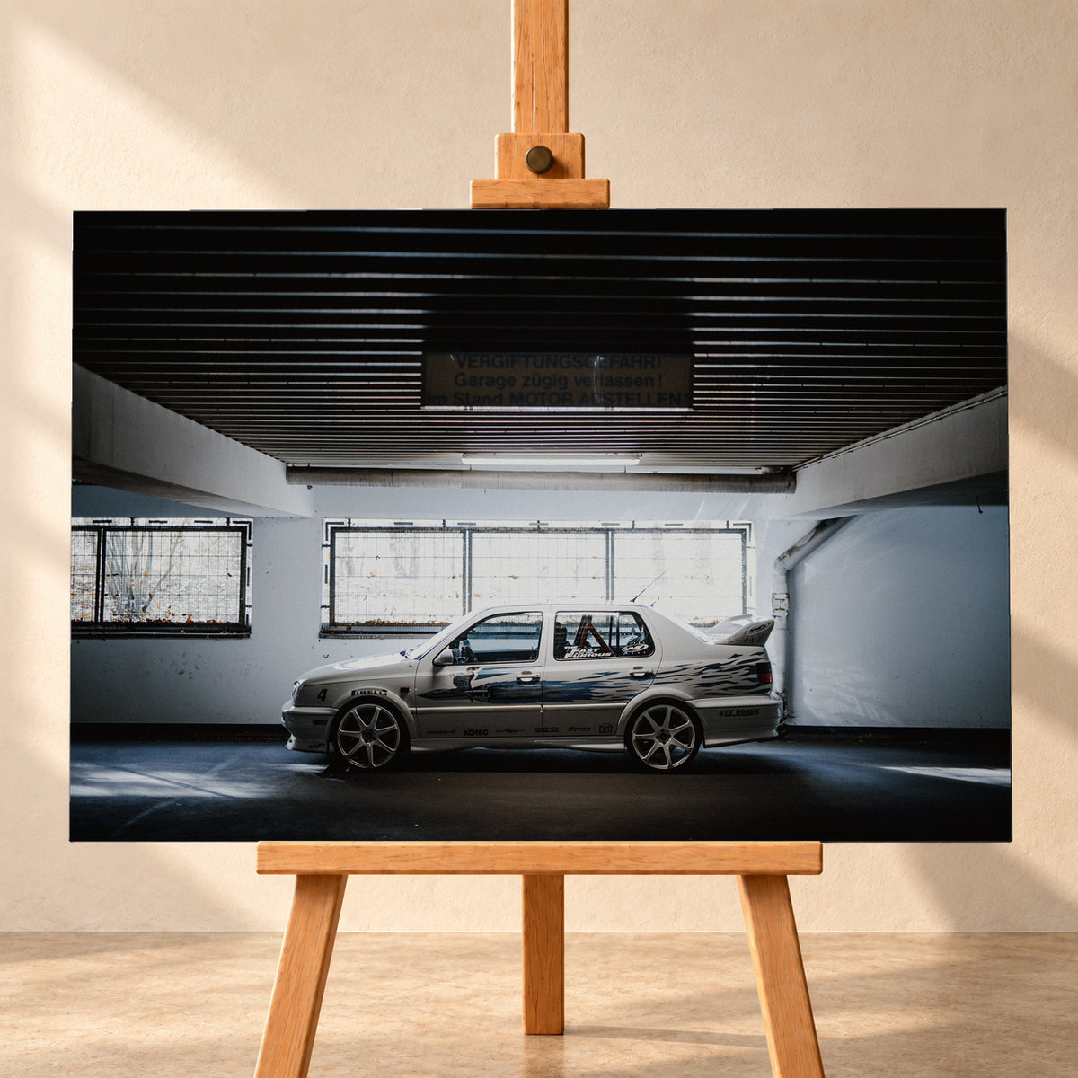 Silberfarbener VW Jetta mit blau-schwarzer Seitengrafik, großen Leichtmetallrädern und hohem Heckflügel im Seitenprofil in einer dunklen Parkgarage; Visualisierung als Leinwand-Wandbild, frontal auf einer Holzstaffelei.