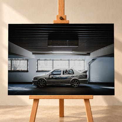 Silberfarbener VW Jetta mit blau-schwarzer Seitengrafik, großen Leichtmetallrädern und hohem Heckflügel im Seitenprofil in einer dunklen Parkgarage; Visualisierung als Leinwand-Wandbild, frontal auf einer Holzstaffelei.