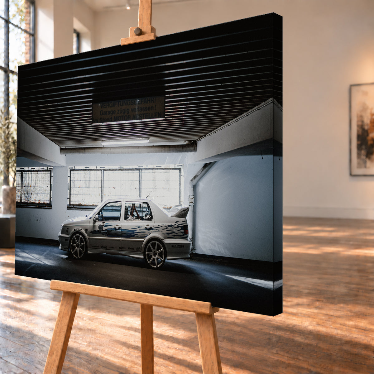 Silberfarbener VW Jetta mit blau-schwarzer Seitengrafik, großen Leichtmetallrädern und hohem Heckflügel im Seitenprofil in einer dunklen Parkgarage; Visualisierung als Leinwand-Wandbild, schräg von der Seite auf einer Holzstaffelei.