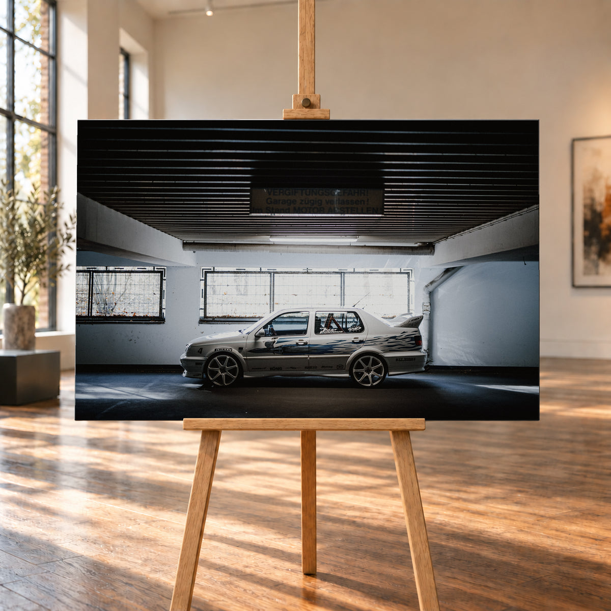 Silberfarbener VW Jetta mit blau-schwarzer Seitengrafik, großen Leichtmetallrädern und hohem Heckflügel im Seitenprofil in einer dunklen Parkgarage; Visualisierung als Leinwand-Wandbild, auf einer Holzstaffelei in einem hellen Galerieraum.