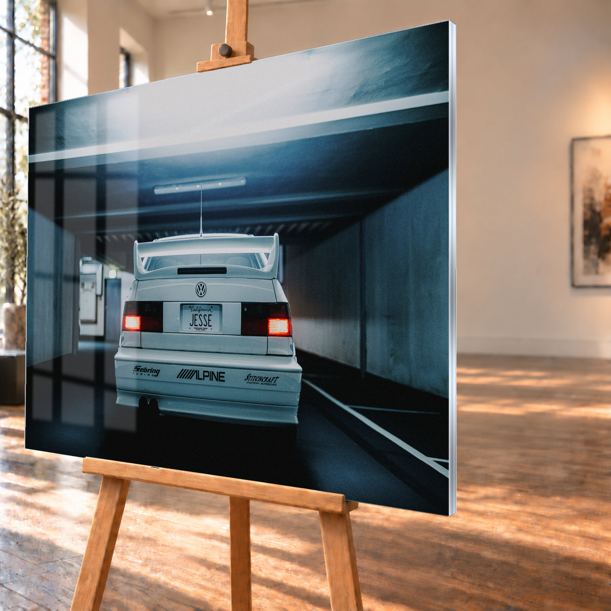 Weißer Volkswagen Jetta in symmetrischer Heckansicht in einer dunklen Betongarage, mit großem Heckflügel, roten Rücklichtern, sichtbaren Aufklebern und kühlem blau-grauem Licht; Visualisierung als Acrylglas-Wandbild, schräg von der Seite auf einer Holzstaffelei.