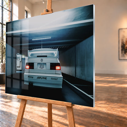 Weißer Volkswagen Jetta in symmetrischer Heckansicht in einer dunklen Betongarage, mit großem Heckflügel, roten Rücklichtern, sichtbaren Aufklebern und kühlem blau-grauem Licht; Visualisierung als Acrylglas-Wandbild, schräg von der Seite auf einer Holzstaffelei.