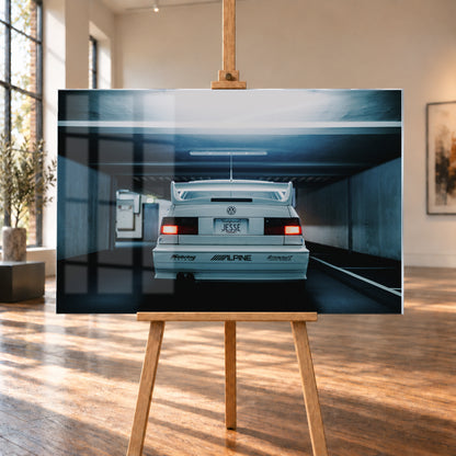 Weißer Volkswagen Jetta in symmetrischer Heckansicht in einer dunklen Betongarage, mit großem Heckflügel, roten Rücklichtern, sichtbaren Aufklebern und kühlem blau-grauem Licht; Visualisierung als Acrylglas-Wandbild, auf einer Holzstaffelei in einem hellen Galerieraum.