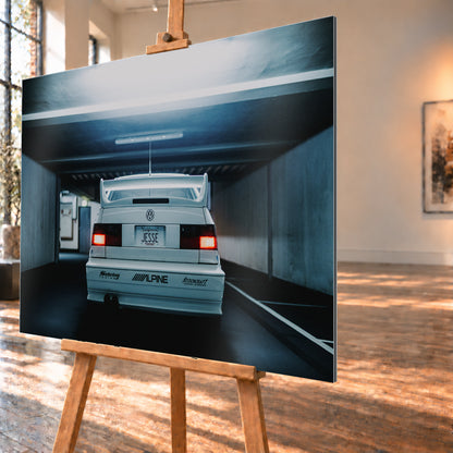 Weißer Volkswagen Jetta in symmetrischer Heckansicht in einer dunklen Betongarage, mit großem Heckflügel, roten Rücklichtern, sichtbaren Aufklebern und kühlem blau-grauem Licht; Visualisierung als Alu gebürstet-Wandbild, schräg von der Seite auf einer Holzstaffelei.