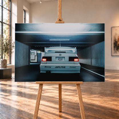 Weißer Volkswagen Jetta in symmetrischer Heckansicht in einer dunklen Betongarage, mit großem Heckflügel, roten Rücklichtern, sichtbaren Aufklebern und kühlem blau-grauem Licht; Visualisierung als Alu gebürstet-Wandbild, auf einer Holzstaffelei in einem hellen Galerieraum.