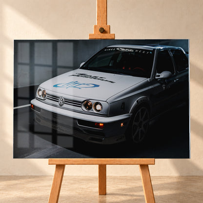 Heller Volkswagen Jetta in frontaler Dreiviertelansicht vor dunklem Hintergrund, mit eingeschalteten runden Frontscheinwerfern, orangefarbenen Leuchten, schwarzen und blauen Grafiken auf der Motorhaube sowie sichtbaren Aufklebern an Scheibe und Karosserie; Visualisierung als Acrylglas-Wandbild, frontal auf einer Holzstaffelei.