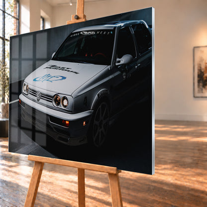 Heller Volkswagen Jetta in frontaler Dreiviertelansicht vor dunklem Hintergrund, mit eingeschalteten runden Frontscheinwerfern, orangefarbenen Leuchten, schwarzen und blauen Grafiken auf der Motorhaube sowie sichtbaren Aufklebern an Scheibe und Karosserie; Visualisierung als Acrylglas-Wandbild, schräg von der Seite auf einer Holzstaffelei.