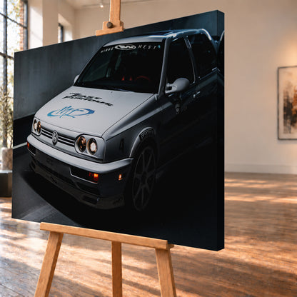 Heller Volkswagen Jetta in frontaler Dreiviertelansicht vor dunklem Hintergrund, mit eingeschalteten runden Frontscheinwerfern, orangefarbenen Leuchten, schwarzen und blauen Grafiken auf der Motorhaube sowie sichtbaren Aufklebern an Scheibe und Karosserie; Visualisierung als Leinwand-Wandbild, schräg von der Seite auf einer Holzstaffelei.
