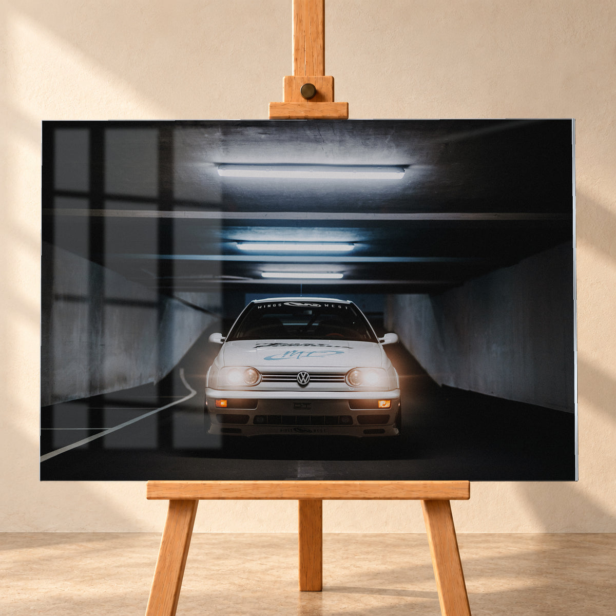 Weißer Volkswagen Jetta in frontaler Ansicht in einer dunklen Betongarage; Scheinwerfer und orangefarbene untere Leuchten sind eingeschaltet, darüber verlaufen helle Deckenröhren; Visualisierung als Acrylglas-Wandbild, frontal auf einer Holzstaffelei.