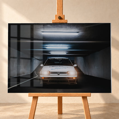 Weißer Volkswagen Jetta in frontaler Ansicht in einer dunklen Betongarage; Scheinwerfer und orangefarbene untere Leuchten sind eingeschaltet, darüber verlaufen helle Deckenröhren; Visualisierung als Acrylglas-Wandbild, frontal auf einer Holzstaffelei.