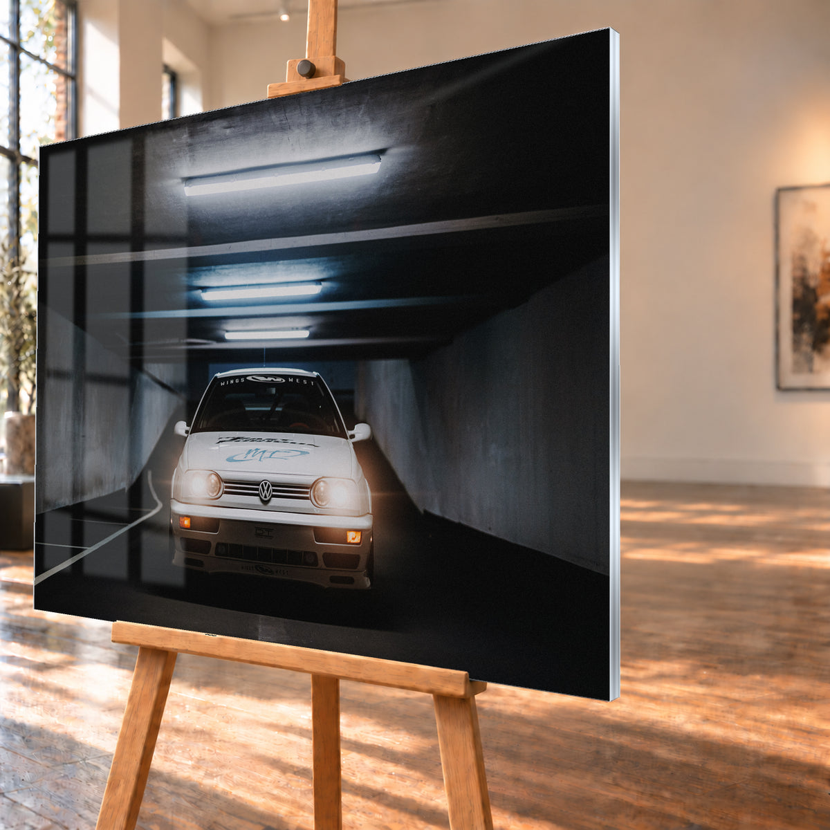 Weißer Volkswagen Jetta in frontaler Ansicht in einer dunklen Betongarage; Scheinwerfer und orangefarbene untere Leuchten sind eingeschaltet, darüber verlaufen helle Deckenröhren; Visualisierung als Acrylglas-Wandbild, schräg von der Seite auf einer Holzstaffelei.