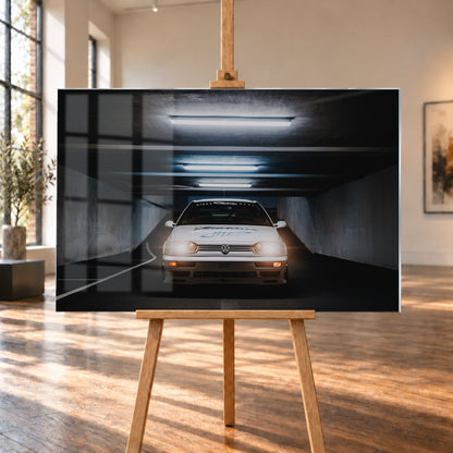 Weißer Volkswagen Jetta in frontaler Ansicht in einer dunklen Betongarage; Scheinwerfer und orangefarbene untere Leuchten sind eingeschaltet, darüber verlaufen helle Deckenröhren; Visualisierung als Acrylglas-Wandbild, auf einer Holzstaffelei in einem hellen Galerieraum.