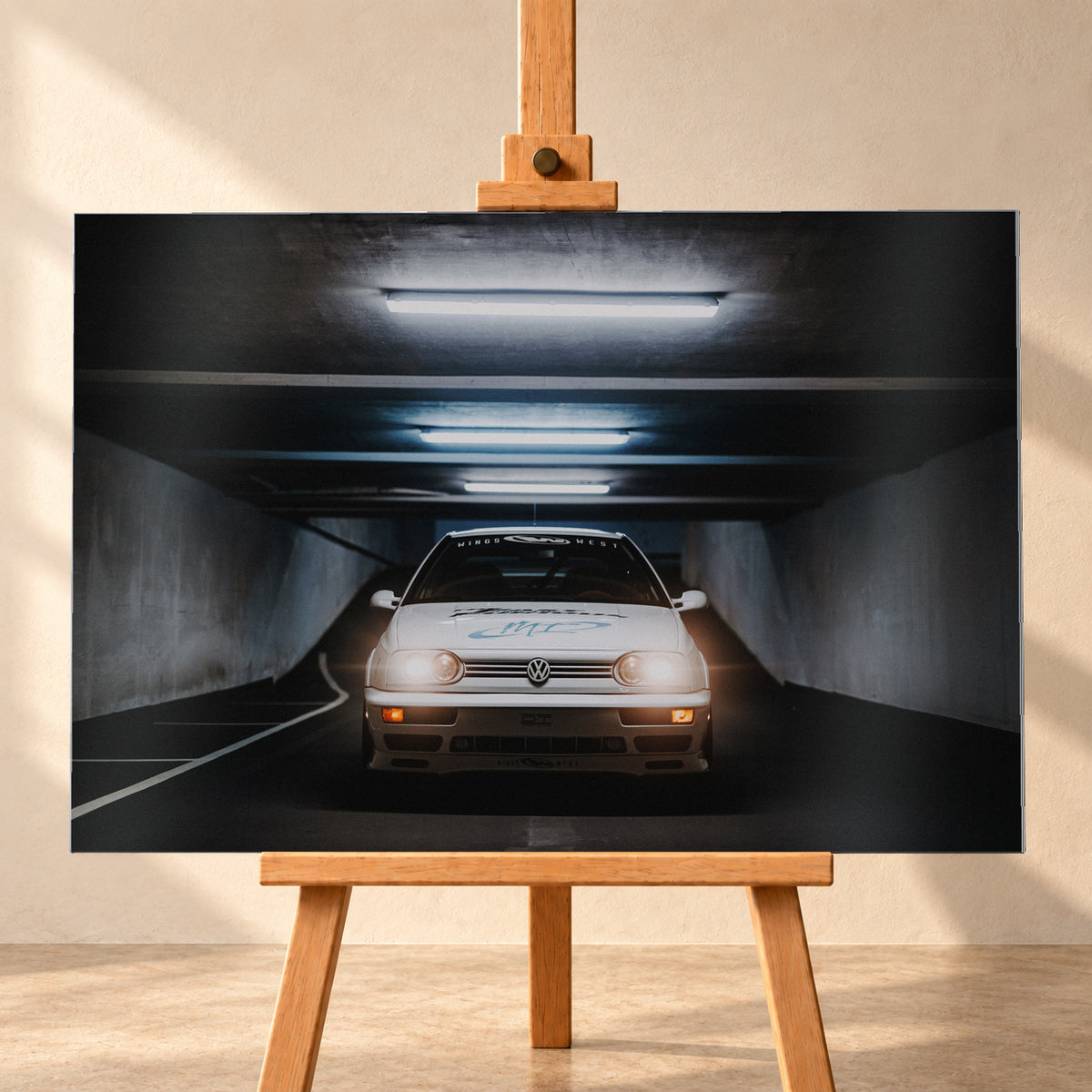 Weißer Volkswagen Jetta in frontaler Ansicht in einer dunklen Betongarage; Scheinwerfer und orangefarbene untere Leuchten sind eingeschaltet, darüber verlaufen helle Deckenröhren; Visualisierung als Alu gebürstet-Wandbild, frontal auf einer Holzstaffelei.