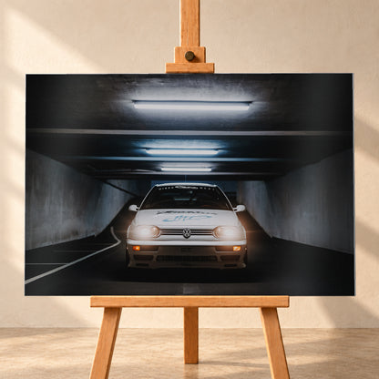 Weißer Volkswagen Jetta in frontaler Ansicht in einer dunklen Betongarage; Scheinwerfer und orangefarbene untere Leuchten sind eingeschaltet, darüber verlaufen helle Deckenröhren; Visualisierung als Alu gebürstet-Wandbild, frontal auf einer Holzstaffelei.