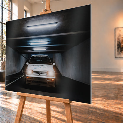 Weißer Volkswagen Jetta in frontaler Ansicht in einer dunklen Betongarage; Scheinwerfer und orangefarbene untere Leuchten sind eingeschaltet, darüber verlaufen helle Deckenröhren; Visualisierung als Alu gebürstet-Wandbild, schräg von der Seite auf einer Holzstaffelei.