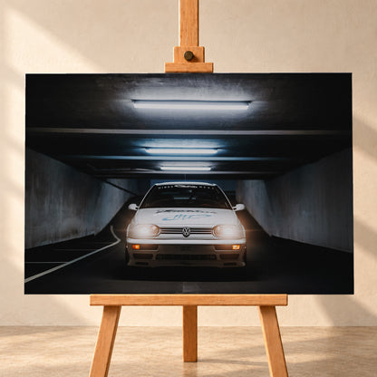 Weißer Volkswagen Jetta in frontaler Ansicht in einer dunklen Betongarage; Scheinwerfer und orangefarbene untere Leuchten sind eingeschaltet, darüber verlaufen helle Deckenröhren; Visualisierung als Leinwand-Wandbild, frontal auf einer Holzstaffelei.