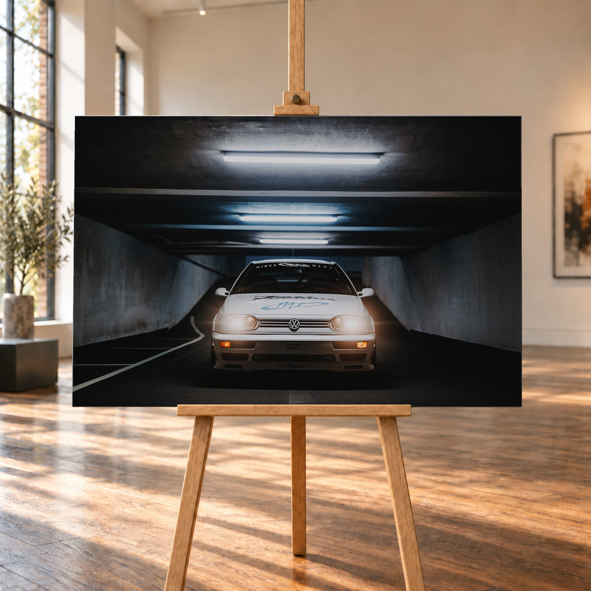 Weißer Volkswagen Jetta in frontaler Ansicht in einer dunklen Betongarage; Scheinwerfer und orangefarbene untere Leuchten sind eingeschaltet, darüber verlaufen helle Deckenröhren; Visualisierung als Leinwand-Wandbild, auf einer Holzstaffelei in einem hellen Galerieraum.