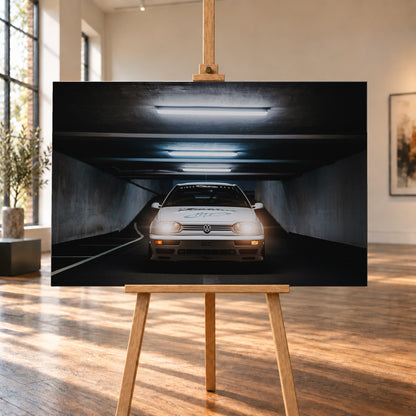 Weißer Volkswagen Jetta in frontaler Ansicht in einer dunklen Betongarage; Scheinwerfer und orangefarbene untere Leuchten sind eingeschaltet, darüber verlaufen helle Deckenröhren; Visualisierung als Leinwand-Wandbild, auf einer Holzstaffelei in einem hellen Galerieraum.