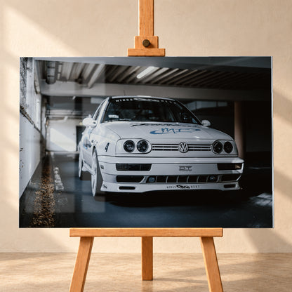 Weißer VW Jetta in tiefer, leicht seitlicher Frontansicht in einer dunklen Parkgarage. Vier runde Scheinwerfer, weiße Räder, Karosseriegrafiken und die Nummer 4 sind sichtbar; links fällt ein heller Lichtkeil auf die Betonwand und die Fahrzeugseite; Visualisierung als Acrylglas-Wandbild, frontal auf einer Holzstaffelei.