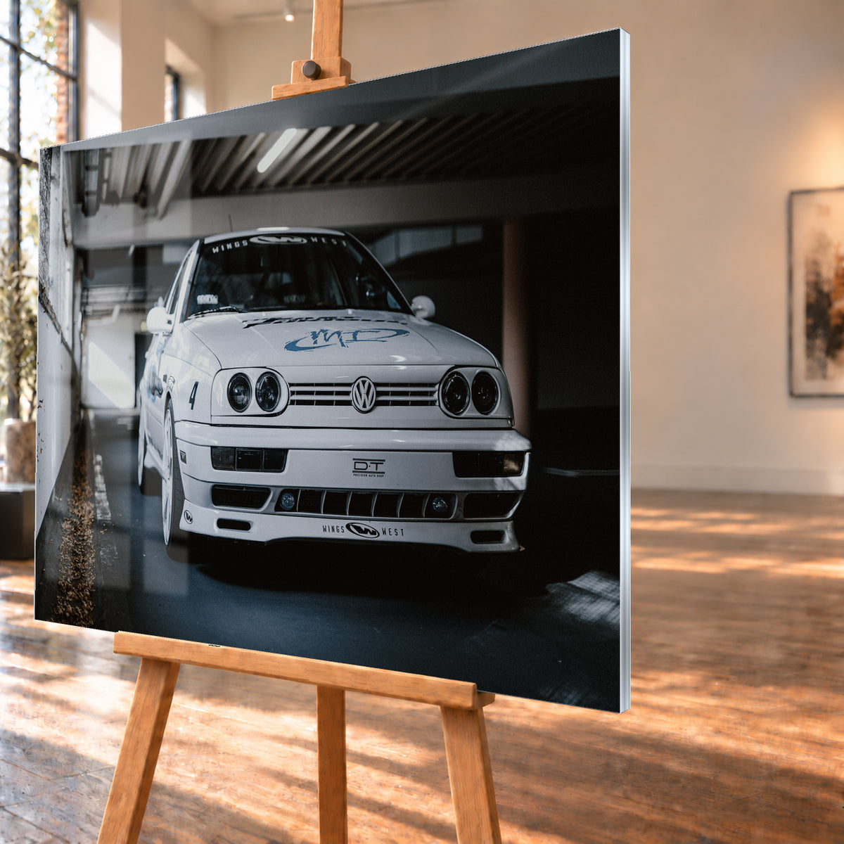 Weißer VW Jetta in tiefer, leicht seitlicher Frontansicht in einer dunklen Parkgarage. Vier runde Scheinwerfer, weiße Räder, Karosseriegrafiken und die Nummer 4 sind sichtbar; links fällt ein heller Lichtkeil auf die Betonwand und die Fahrzeugseite; Visualisierung als Acrylglas-Wandbild, schräg von der Seite auf einer Holzstaffelei.