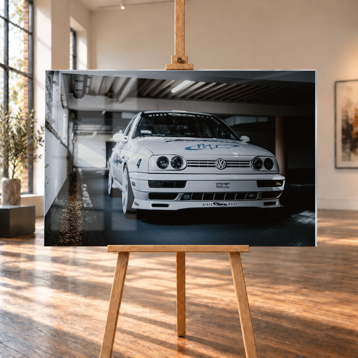 Weißer VW Jetta in tiefer, leicht seitlicher Frontansicht in einer dunklen Parkgarage. Vier runde Scheinwerfer, weiße Räder, Karosseriegrafiken und die Nummer 4 sind sichtbar; links fällt ein heller Lichtkeil auf die Betonwand und die Fahrzeugseite; Visualisierung als Acrylglas-Wandbild, auf einer Holzstaffelei in einem hellen Galerieraum.