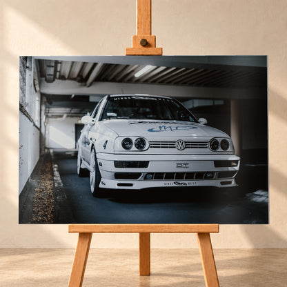 Weißer VW Jetta in tiefer, leicht seitlicher Frontansicht in einer dunklen Parkgarage. Vier runde Scheinwerfer, weiße Räder, Karosseriegrafiken und die Nummer 4 sind sichtbar; links fällt ein heller Lichtkeil auf die Betonwand und die Fahrzeugseite; Visualisierung als Alu gebürstet-Wandbild, frontal auf einer Holzstaffelei.
