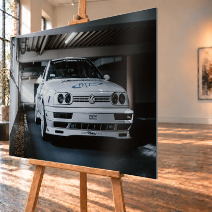 Weißer VW Jetta in tiefer, leicht seitlicher Frontansicht in einer dunklen Parkgarage. Vier runde Scheinwerfer, weiße Räder, Karosseriegrafiken und die Nummer 4 sind sichtbar; links fällt ein heller Lichtkeil auf die Betonwand und die Fahrzeugseite; Visualisierung als Alu gebürstet-Wandbild, schräg von der Seite auf einer Holzstaffelei.