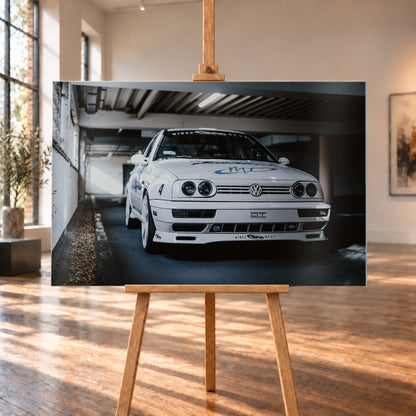 Weißer VW Jetta in tiefer, leicht seitlicher Frontansicht in einer dunklen Parkgarage. Vier runde Scheinwerfer, weiße Räder, Karosseriegrafiken und die Nummer 4 sind sichtbar; links fällt ein heller Lichtkeil auf die Betonwand und die Fahrzeugseite; Visualisierung als Alu gebürstet-Wandbild, auf einer Holzstaffelei in einem hellen Galerieraum.