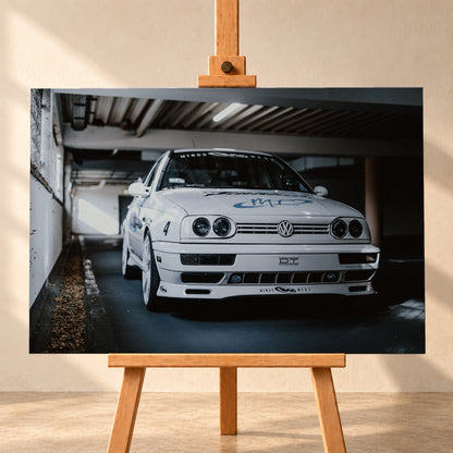 Weißer VW Jetta in tiefer, leicht seitlicher Frontansicht in einer dunklen Parkgarage. Vier runde Scheinwerfer, weiße Räder, Karosseriegrafiken und die Nummer 4 sind sichtbar; links fällt ein heller Lichtkeil auf die Betonwand und die Fahrzeugseite; Visualisierung als Leinwand-Wandbild, frontal auf einer Holzstaffelei.