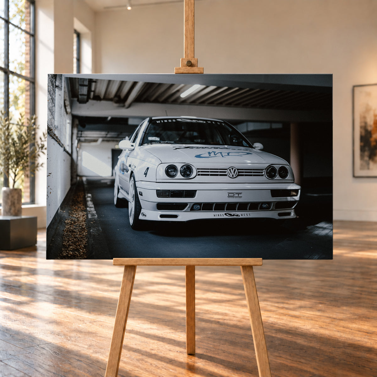 Weißer VW Jetta in tiefer, leicht seitlicher Frontansicht in einer dunklen Parkgarage. Vier runde Scheinwerfer, weiße Räder, Karosseriegrafiken und die Nummer 4 sind sichtbar; links fällt ein heller Lichtkeil auf die Betonwand und die Fahrzeugseite; Visualisierung als Leinwand-Wandbild, auf einer Holzstaffelei in einem hellen Galerieraum.