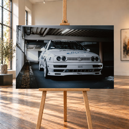 Weißer VW Jetta in tiefer, leicht seitlicher Frontansicht in einer dunklen Parkgarage. Vier runde Scheinwerfer, weiße Räder, Karosseriegrafiken und die Nummer 4 sind sichtbar; links fällt ein heller Lichtkeil auf die Betonwand und die Fahrzeugseite; Visualisierung als Leinwand-Wandbild, auf einer Holzstaffelei in einem hellen Galerieraum.