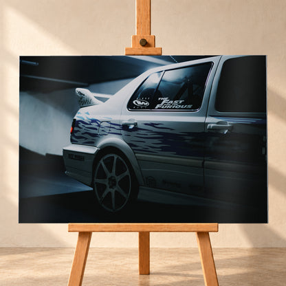 Nahaufnahme der hinteren Seitenpartie eines hellen VW Jetta mit blau-violetter Flammengrafik, großem Rad und Heckflügel. Im dunklen Seitenfenster steht „The Fast and the Furious“; kühles Licht fällt auf Wand und Boden; Visualisierung als Alu gebürstet-Wandbild, frontal auf einer Holzstaffelei.