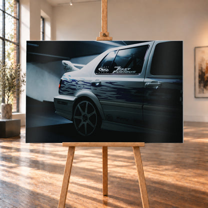 Nahaufnahme der hinteren Seitenpartie eines hellen VW Jetta mit blau-violetter Flammengrafik, großem Rad und Heckflügel. Im dunklen Seitenfenster steht „The Fast and the Furious“; kühles Licht fällt auf Wand und Boden; Visualisierung als Alu gebürstet-Wandbild, auf einer Holzstaffelei in einem hellen Galerieraum.