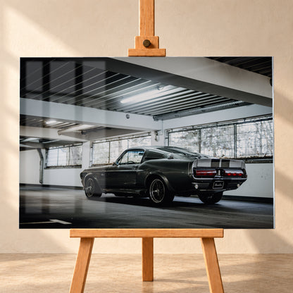 Dunkelgraue Eleanor mit hellen Doppelstreifen in tiefer Heck-Dreiviertelansicht auf einer leeren Parkebene mit weißen Trägern, großen Fenstern und kühlem Deckenlicht; Visualisierung als Acrylglas-Wandbild, frontal auf einer Holzstaffelei.