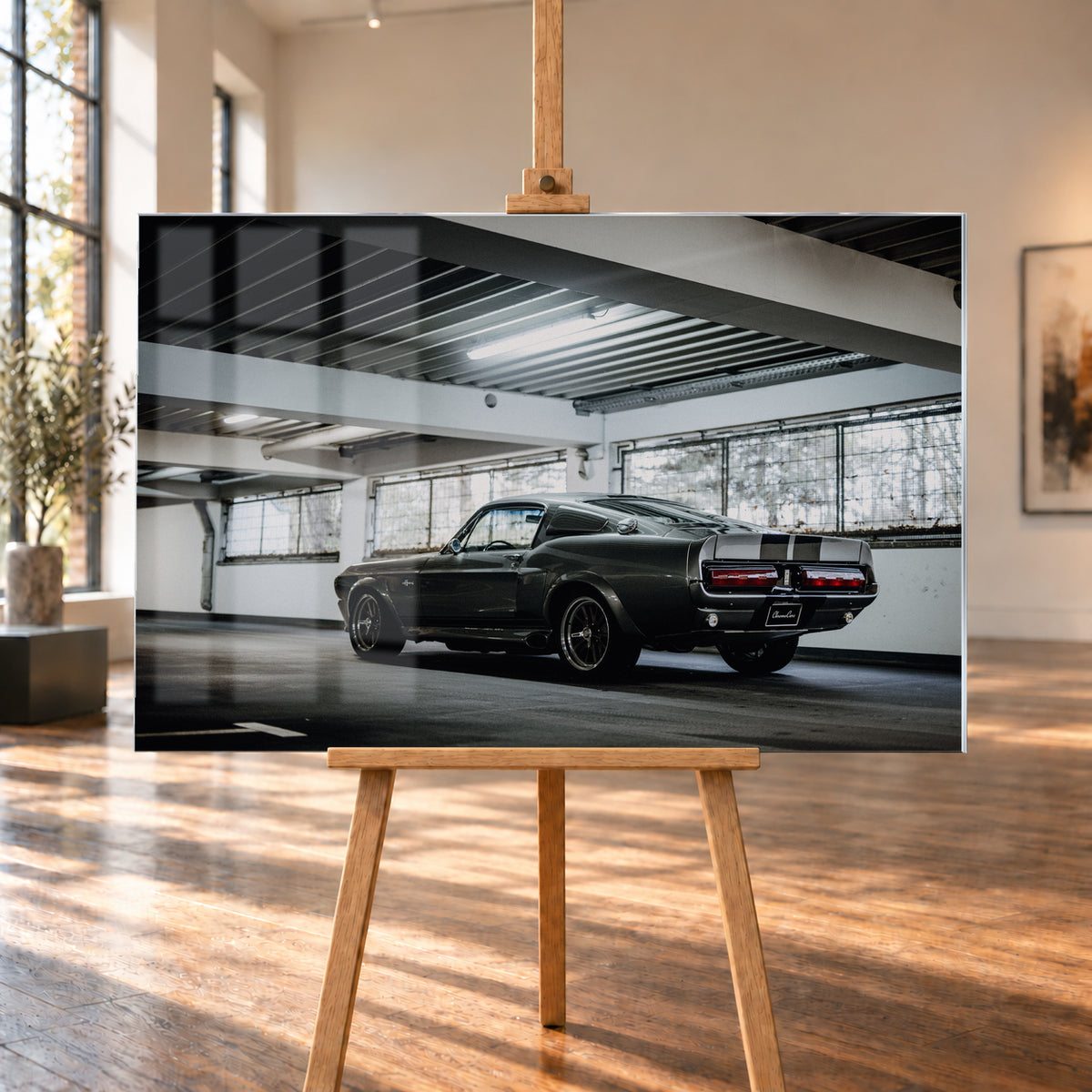 Dunkelgraue Eleanor mit hellen Doppelstreifen in tiefer Heck-Dreiviertelansicht auf einer leeren Parkebene mit weißen Trägern, großen Fenstern und kühlem Deckenlicht; Visualisierung als Acrylglas-Wandbild, auf einer Holzstaffelei in einem hellen Galerieraum.