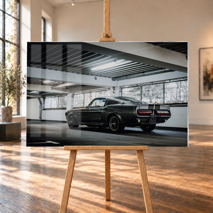 Dunkelgraue Eleanor mit hellen Doppelstreifen in tiefer Heck-Dreiviertelansicht auf einer leeren Parkebene mit weißen Trägern, großen Fenstern und kühlem Deckenlicht; Visualisierung als Acrylglas-Wandbild, auf einer Holzstaffelei in einem hellen Galerieraum.