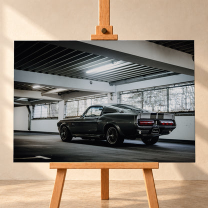 Dunkelgraue Eleanor mit hellen Doppelstreifen in tiefer Heck-Dreiviertelansicht auf einer leeren Parkebene mit weißen Trägern, großen Fenstern und kühlem Deckenlicht; Visualisierung als Leinwand-Wandbild, frontal auf einer Holzstaffelei.
