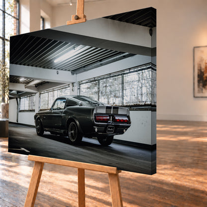 Dunkelgraue Eleanor mit hellen Doppelstreifen in tiefer Heck-Dreiviertelansicht auf einer leeren Parkebene mit weißen Trägern, großen Fenstern und kühlem Deckenlicht; Visualisierung als Leinwand-Wandbild, schräg von der Seite auf einer Holzstaffelei.