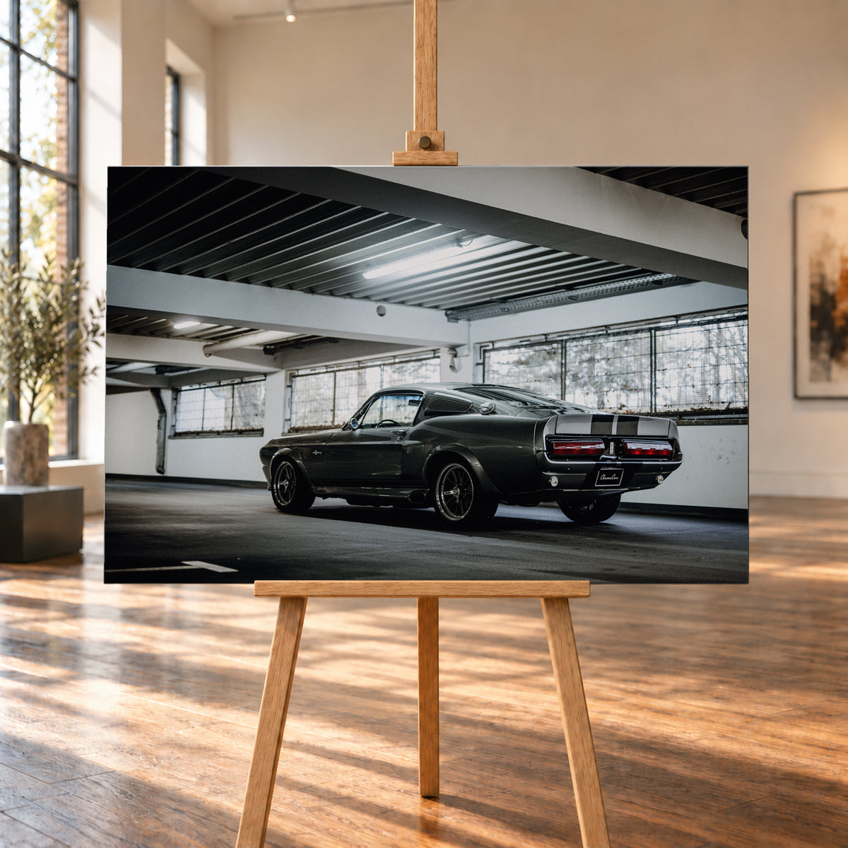 Dunkelgraue Eleanor mit hellen Doppelstreifen in tiefer Heck-Dreiviertelansicht auf einer leeren Parkebene mit weißen Trägern, großen Fenstern und kühlem Deckenlicht; Visualisierung als Leinwand-Wandbild, auf einer Holzstaffelei in einem hellen Galerieraum.